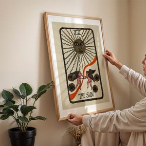 May include: A framed art print of The Sun tarot card. The artwork features a central sun design with radiating lines, a border, and the text "XIX" at the top. Two figures are depicted holding hands, with the text "THE SUN" at the bottom.