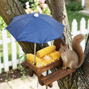 May include: A wooden picnic table bird feeder with a blue parasol, filled with corn on the cob. A squirrel sits on the table. The feeder is attached to a tree branch, with a white picket fence in the background. The scene is set outdoors.