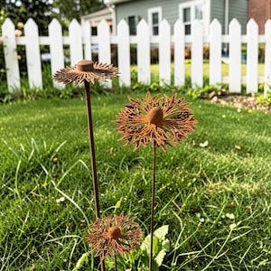 May include: Three rusty metal flower garden stakes with intricate cut-out designs. The flowers are in varying sizes and heights, set against a backdrop of green grass and a white picket fence. Decorative outdoor garden art.