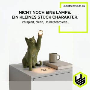 Op de afbeelding: Groene katvormige lamp met een opgeheven poot die licht uitstraalt. De lamp staat op een wit oppervlak met een mok, boek en pootafdruk. Tekst op de afbeelding luidt "NICHT NOCH EINE LAMPE. EIN KLEINES STÜCK CHARAKTER."