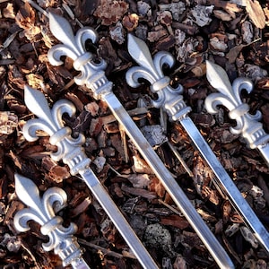 May include: Five silver metal skewers with fleur-de-lis shaped handles. The skewers are lying on a bed of brown wood chips.