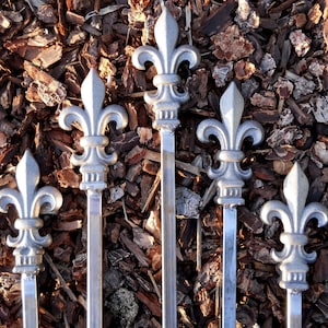 May include: Five silver metal skewers with fleur-de-lis shaped tops. The skewers are arranged in a fan shape against a background of brown wood chips.