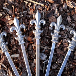 May include: Five silver metal fleur-de-lis finials with long, slender stems. The finials are arranged in a fan shape on a bed of brown wood chips.