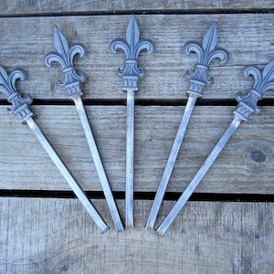 May include: Five silver-colored metal fleur-de-lis finials with pointed tips. The finials are arranged in a fan shape on a wooden surface.