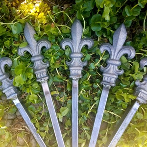 May include: Six silver metal fleur-de-lis finials with pointed tips, arranged in a fan shape on a bed of green foliage.