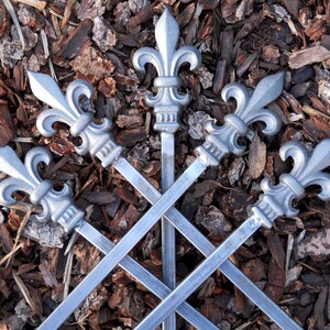 May include: Five silver metal skewers with fleur-de-lis shaped handles. The skewers are crossed over each other and are lying on a bed of brown wood chips.