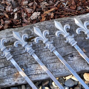 May include: Six silver metal skewers with fleur-de-lis shaped handles. The skewers are arranged in a row on a wooden surface.