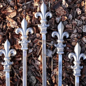 May include: Five silver metal fleur-de-lis finials with a pointed tip. The finials are arranged vertically and are set against a background of brown wood chips.