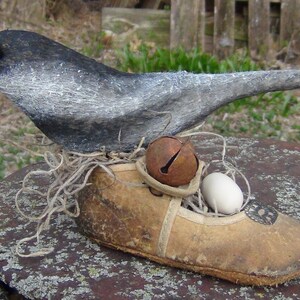 May include: A gray and white bird figurine perched on a brown leather shoe with a white egg and a rusty bell. The bird is made of fabric and has a black eye. The shoe is decorated with a brown leather strap and a metal buckle.