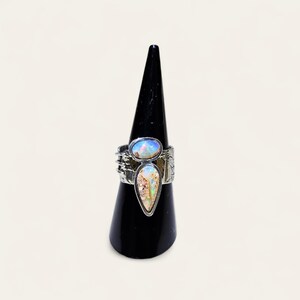 May include: A silver ring with two opal stones. The top stone is oval-shaped and the bottom stone is teardrop-shaped. The ring has a textured band with etched details. The opals have a mix of colors, including blue, white, and brown.