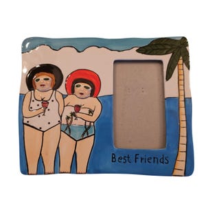 May include: A ceramic picture frame with a beach theme. Two figures in swimsuits and hats are holding wine glasses. The frame has a blue and white background with a palm tree and the words "Best Friends".