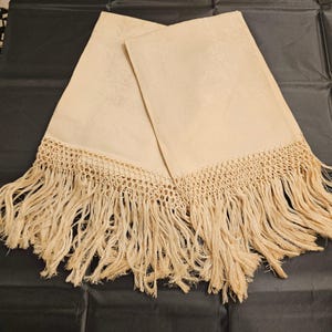 May include: Two rectangular cream-colored fabric towels featuring a subtle tonal damask pattern. Each towel is finished with a wide, intricate crocheted border and long, loose fringe along the bottom edge, creating a textured appearance.