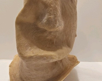 Könnte beinhalten: Eine hellbraune Skulptur eines Moai-Kopfes mit markanter Nase, geschlossenem Mund und leicht geneigtem Kopf. Die Skulptur hat eine strukturierte Oberfläche und steht auf einem Sockel.