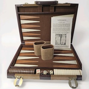 May include: A brown briefcase-style backgammon set featuring a felt interior with tan and brown triangular points. The set includes two tan dice cups, a set of brown and white checkers, dice, a doubling cube, and a printed Backgammon Player's Guide.