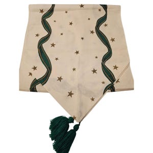 May include: Cream-coloured table runner featuring gold stars and green ribbon patterns. The runner has two green tassels at the bottom. A decorative piece for a dining table or other surface.