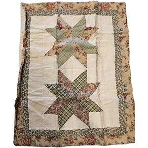 Op de afbeelding: Een rechthoekige quilt met twee grote sterpatronen in het midden op een crèmekleurige achtergrond. De sterren zijn gemaakt van groen geruite en bloemrijke stof. De quilt is omrand met een donkere bloemenprint en een brede bloemrijke rand.