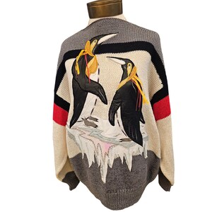 Peut inclure: Un pull en tricot à manches longues avec un panneau central crème orné de deux appliqués de manchots noirs et blancs sur un bloc de glace. Le pull présente des rayures horizontales noires, rouges et grises, avec un col rond côtelé.