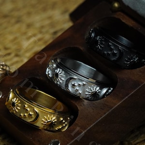 Puede incluir: Tres anillos personalizados con un diseño de sol y luna. Los anillos se muestran en una caja de madera con forro de terciopelo. Los anillos están hechos de diferentes metales, incluyendo oro, plata y negro.