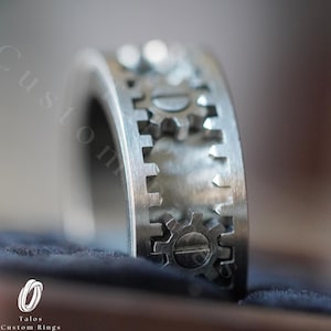 May include: A silver ring with a design of interlocking gears. The ring is made of metal and has a brushed finish.