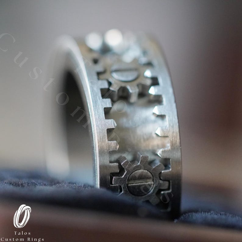 May include: A silver ring with a design of interlocking gears. The ring is made of metal and has a brushed finish.
