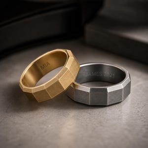 May include: Two faceted rings, one gold and one silver, rest on a gray surface. The gold ring has "MIA" engraved inside. The silver ring has "JAMES 2021" engraved inside. Both rings have a geometric design.