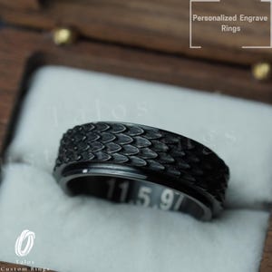 以下が含まれることがあります： 鱗模様のテクスチャが施された黒いリング。リングには「11.5.97」という白い刻印があります。