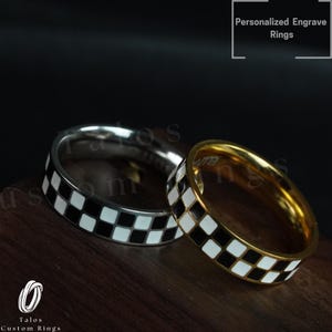 Könnte beinhalten: Zwei Ringe mit einem schwarz-weißen Karomuster. Ein Ring ist silber und der andere ist gold. Die Ringe liegen auf einer Holzoberfläche. Personalisierte Gravurringe.