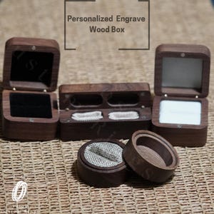 May include: Four wooden ring boxes, two with a single compartment and two with two compartments. The boxes are made of dark wood and have a natural finish. The boxes are open and show the interior lining, which is made of a soft, white fabric. The boxes are arranged on a woven surface.