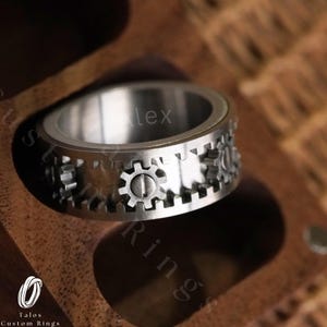 May include: A silver spinning ring with a gear design. The ring is in a wooden box.