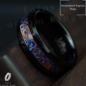 Puede incluir: Anillo de carburo de tungsteno negro con una incrustación azul y oro rosa de engranajes. El anillo está grabado con el nombre "John".