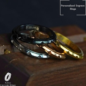 May include: Three stacked rings with a textured, triangular edge. The top ring is black and engraved with the name "Emma". The middle ring is silver and the bottom ring is gold.