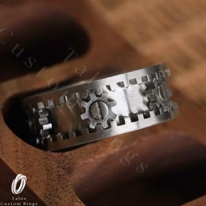 May include: A silver ring with a gear design. The ring is made of metal and has a polished finish.
