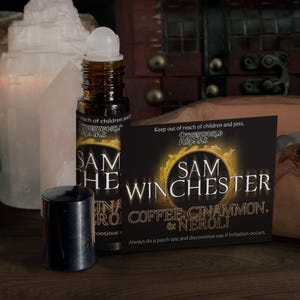 May include: A brown glass roll-on bottle with a black cap, a product card, and a white crystal formation. The product card reads "SAM WINCHESTER COFFEE, CINNAMON, & NEROLI". The bottle is labeled "SAM THE INA RO".