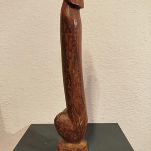 May include: A carved wooden sculpture of a phallus, standing upright. The sculpture is a dark brown color and appears to be made from a single piece of wood. The base is a rough, natural shape, and the overall form is smooth and rounded.