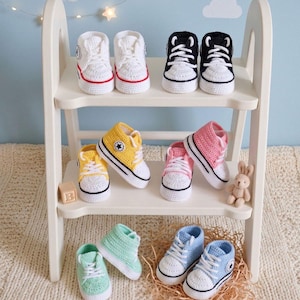 May include: A collection of handmade crochet baby sneakers in various colors including white, black, yellow, pink, mint green, and light blue. Each pair features white laces, a round toe cap, and a small circular patch on the side of the ankle.