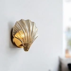 Könnte beinhalten: Eine goldene Wandleuchte mit einem muschelförmigen Glasschirm. Der Schirm ist hellbraun und hat eine geriffelte Textur.