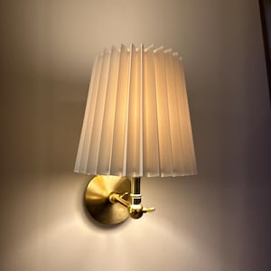 Puede incluir: Una lámpara de pared con una pantalla plisada. La pantalla es de color beige claro y está hecha de una tela texturizada. La lámpara tiene una base y un brazo de color latón. La luz es cálida y acogedora.