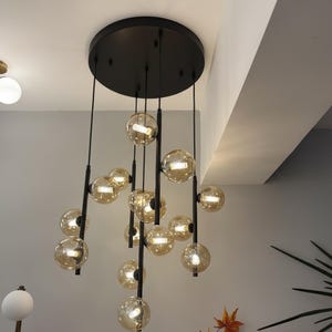 May include: Contemporary chandelier with numerous amber glass spheres hanging from a black ceiling fitting. The light fittings are positioned at different levels, producing a dynamic and stylish lighting design.