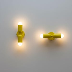 May include: Two bright yellow wall-mounted light fixtures with cylindrical bodies and rounded ends. Each fixture features two glowing white bulbs at the tips, emitting a warm light. One unit is oriented vertically, while the other is positioned horizontally.