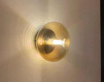 Könnte beinhalten: Eine runde, bernsteinfarbene Glühbirne ist an einer weißen Wand befestigt. Die Glühbirne leuchtet und strahlt ein warmes Licht aus, das ein kreisförmiges Muster um die Leuchte erzeugt. Die Leuchte hat ein schlichtes, modernes Design.