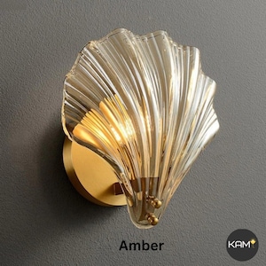Op de afbeelding: Amberkleurige glazen wandlamp in een schelpontwerp. De armatuur heeft een goudkleurige metalen basis en een gegolfde glazen kap. Het woord "Amber" staat onderaan de afbeelding. De lamp is op een grijze muur gemonteerd.