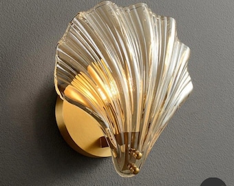 May include: Amber-colored glass wall sconce in a seashell design. The fixture has a gold-colored metal base and a rippled glass shade. The word "Amber" is printed at the bottom of the image. The sconce is mounted on a gray wall.