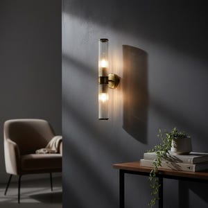 Puede incluir: Aplique de pared moderno con dos tubos de vidrio transparente acanalado, cada uno con una bombilla cálida. La lámpara tiene una base y detalles metálicos de color latón, montada en una pared gris oscuro. Diseño elegante y contemporáneo.