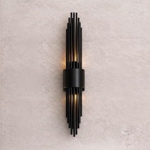 May include: A vertical wall sconce featuring a central matte black cylindrical band surrounded by a series of thin, parallel black metal rods that extend upward and downward, emitting a warm, soft glow from the top and bottom sections.