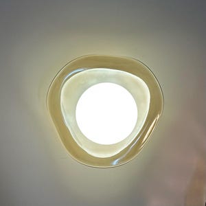 May include: A wall-mounted light fixture featuring a circular, glowing white bulb centered within a translucent, amber-tinted glass shade. The glass shade has an irregular, organic, wavy edge that frames the bright light source.