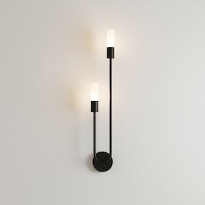 May include: A contemporary black wall sconce featuring two vertical lights. Each light has a cylindrical white shade and a black base. The sconce is mounted on a circular black base, ideal for modern interiors.