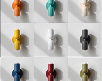 May include: A collection of nine wall-mounted light fixtures, each featuring a circular base and two cylindrical, vertically aligned shades. The lamps are displayed in various solid colors including orange, teal, cream, yellow, white, gray, blue, red, and olive green.