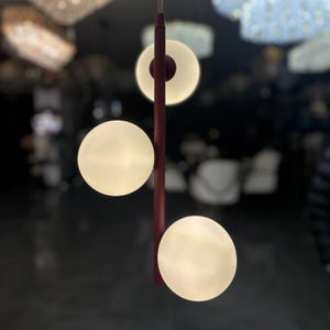 May include: A contemporary pendant light fitting featuring three illuminated orbs and a circular light. The fixture has a burgundy stem and is suspended from the ceiling. The orbs and circular light emit a warm glow, suitable for interior lighting.
