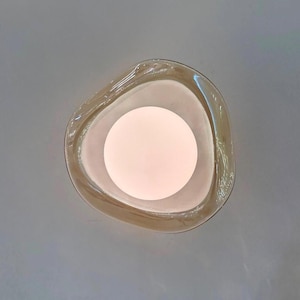 Pode incluir: Uma vista de cima de uma tigela de vidro translúcido tingido de âmbar com uma forma triangular arredondada, orgânica e irregular. O vidro apresenta bordas suaves e curvas e um acabamento polido, contendo uma fonte de luz branca circular.