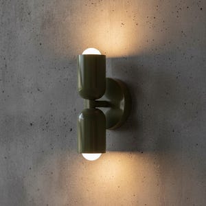 Puede incluir: Un aplique de pared de doble luz con un cuerpo cilíndrico de metal verde oliva y dos extremos redondeados. La lámpara está montada en la pared y emite un brillo cálido y suave desde las aberturas superior e inferior del dispositivo.